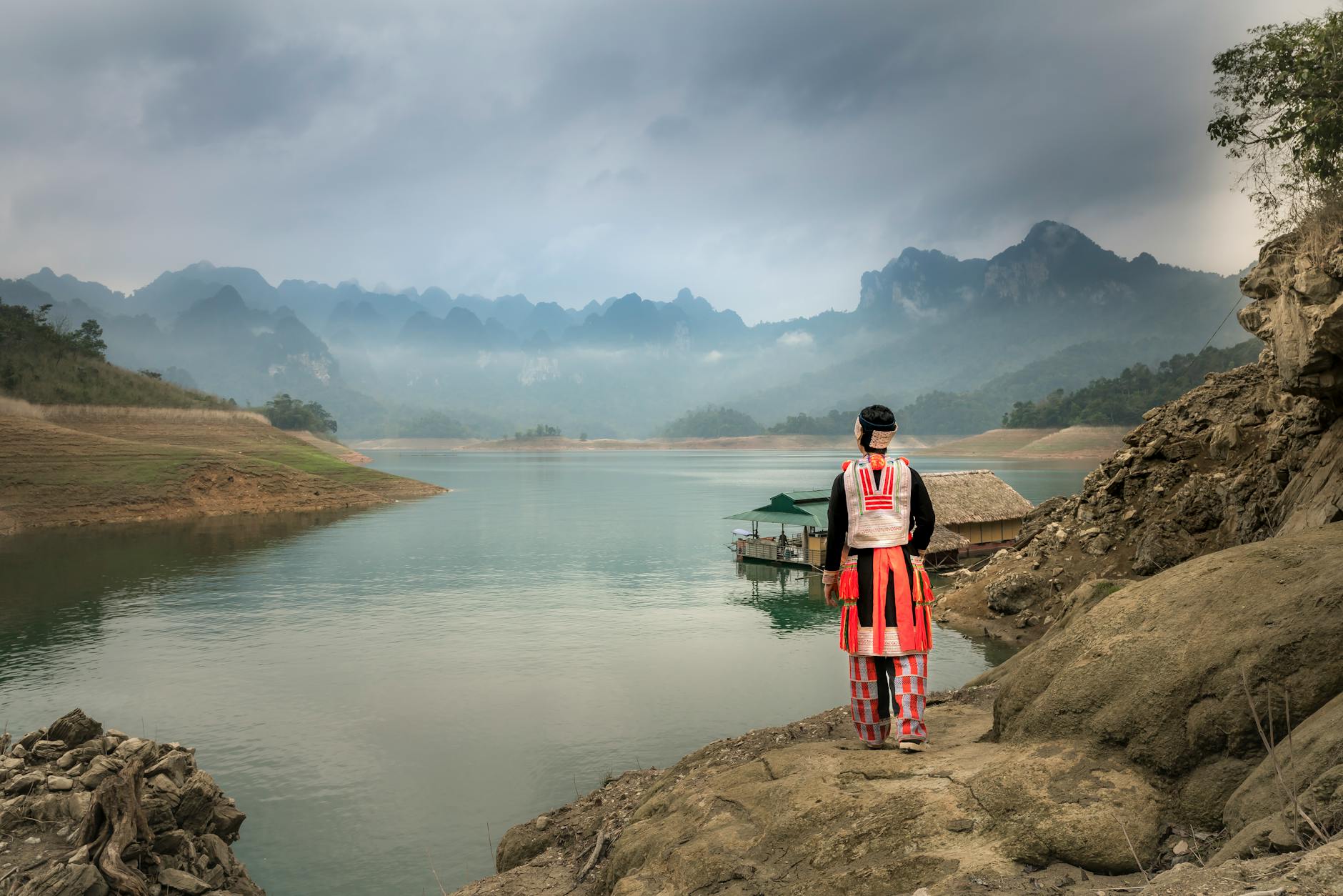 A woman in traditional Thai clothing kneeling by a river, gently releasing a banana-leaf krathong at sunset
